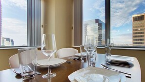 Mesa de comedor para cuatro personas, con vasos, platos y cubiertos limpios, ubicada cerca de amplios ventanales con vista a la ciudad.