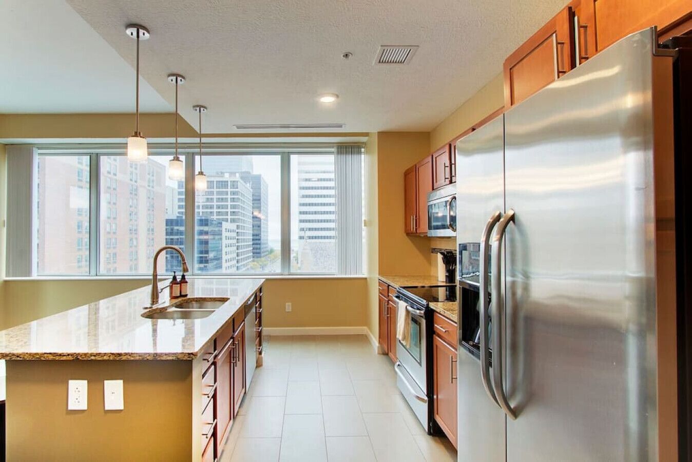 A modern kitchen with a large island, stainless steel appliances, and a view of tall buildings through the window.