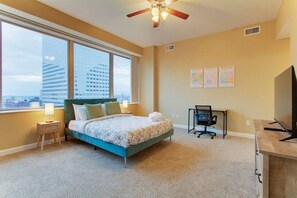 A bedroom with a bed, a desk, a chair, a TV, and large windows with a city view. - Luxury High Rise | Downtown | Free Parking (Cleveland)