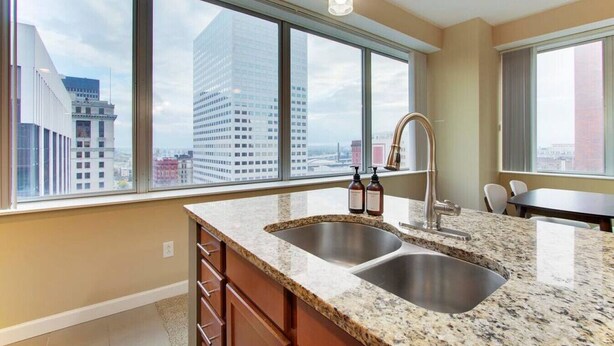 A modern kitchen with a double sink, granite countertop, and a view of a cityscape through large windows. - Luxury High Rise | Downtown | Free Parking (Cleveland)