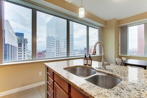 A modern kitchen with a double sink, granite countertop, and a view of a cityscape through large windows. - Luxury High Rise | Downtown | Free Parking (Cleveland)