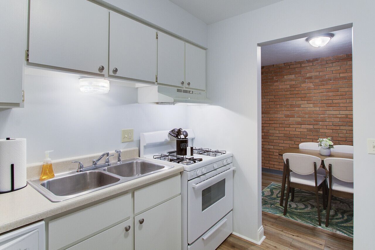 A compact kitchen with white cabinets, a gas stove, a sink, and a dining area with a brick wall and a table.