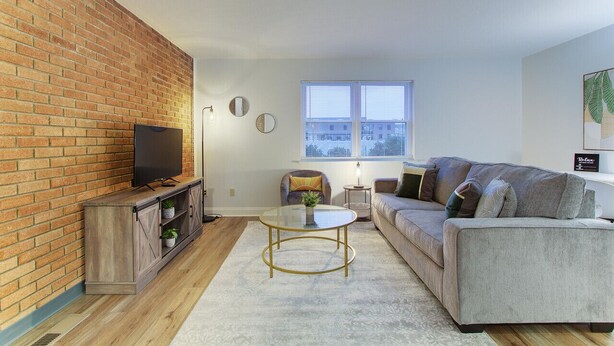 Ein Wohnzimmer mit Backsteinwand, einem Flachbildfernseher auf einer Holzkommode, einem grauen Sofa, einem Glastisch und einem Fenster mit Jalousien.
