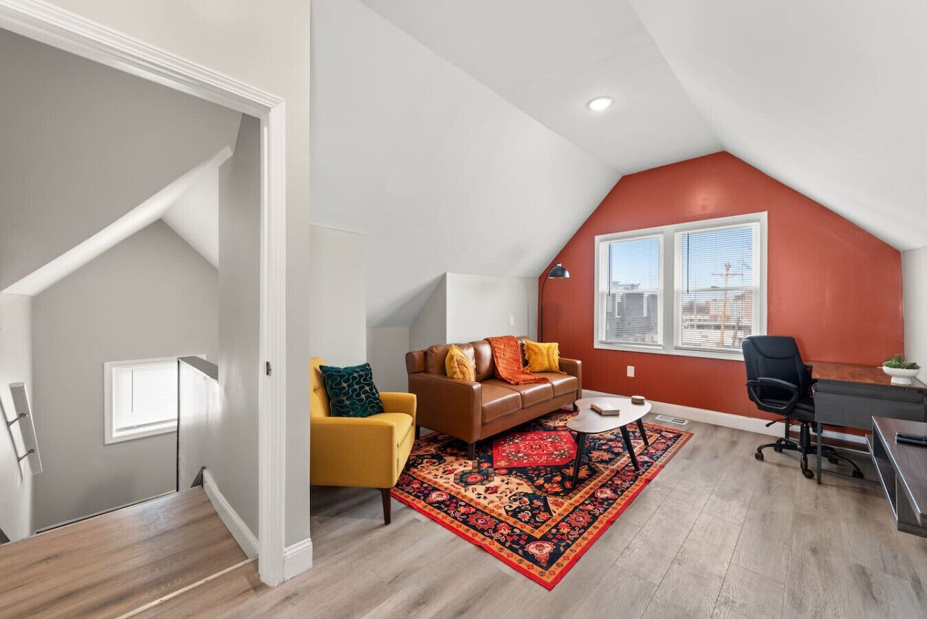 A loft-style room with a sloped ceiling, a brown leather sofa, a yellow armchair, a small round table, and a red patterned rug.