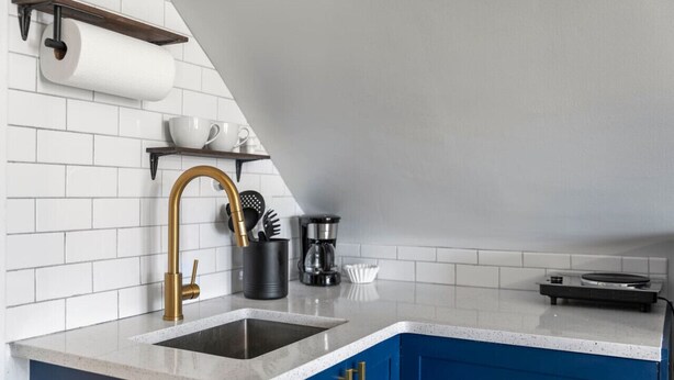Une cuisine avec un mur recouvert de carreaux blancs, un robinet doré, une machine à café et une petite étagère avec des tasses et des ustensiles.