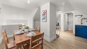 Un coin repas avec une table en bois, des chaises et un vase de fleurs. On aperçoit une cuisine à l’arrière-plan, avec des armoires bleues et des appareils électroménagers blancs.