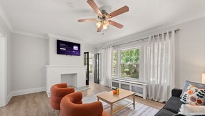 A modern living room with a fireplace, a ceiling fan, a flat-screen TV, and a large window with curtains. - 2BR Near Cleveland Clinic | Garage & Patio (Cleveland Heights)