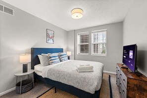 A bedroom with a bed, a nightstand, a lamp, a TV, and a window with blinds.