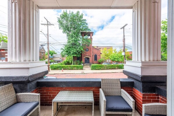 A covered outdoor seating area with wicker furniture and a view of a brick building with a bell tower.