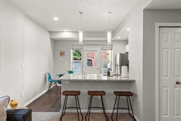 Una cocina moderna con encimera de mármol, taburetes de bar, luces colgantes y vistas al exterior.