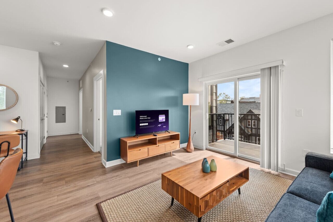 A modern living room with a wooden coffee table, a sofa, a television on a wooden stand, and a large sliding glass door leading to a balcony.