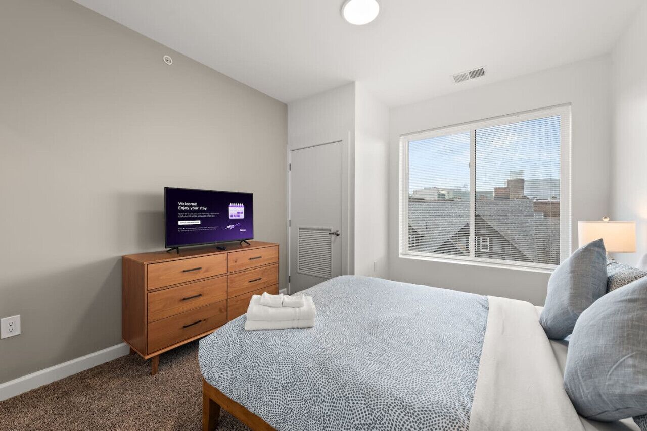 A bedroom with a bed, a TV on a wooden dresser, a window with blinds, and a door.