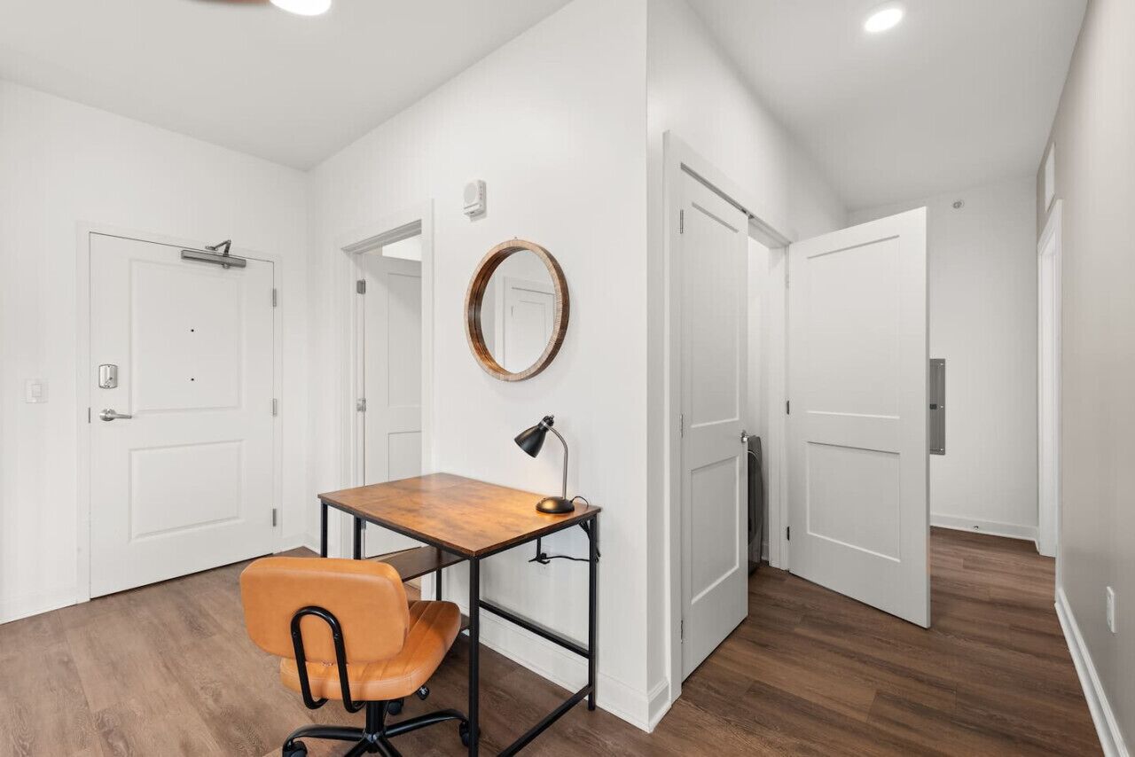 Una habitación con un escritorio de madera, una silla naranja, un espejo redondo y puertas blancas.