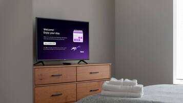 Un televisor mostrando un mensaje de bienvenida, un aparador de madera con cajones y una toalla blanca doblada cuidadosamente sobre una cama.