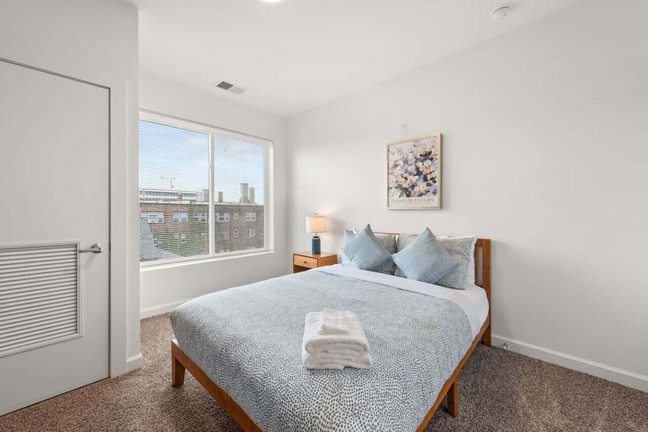 A bedroom with a bed, a window with blinds, a nightstand with a lamp, and a framed picture on the wall.