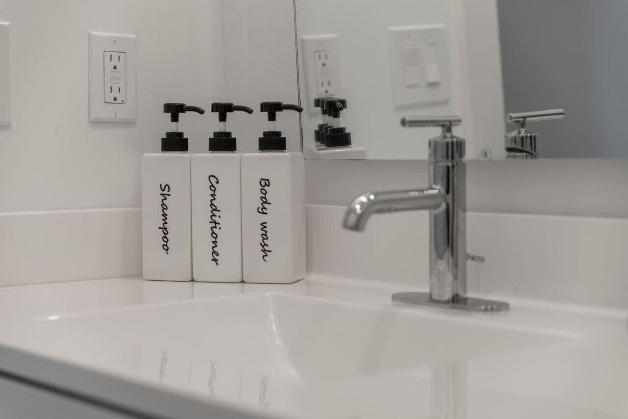 A bathroom with a white sink, a chrome faucet, and three bottles of liquid soap labeled 'Shampoo,' 'Conditioner,' and 'Body wash.'