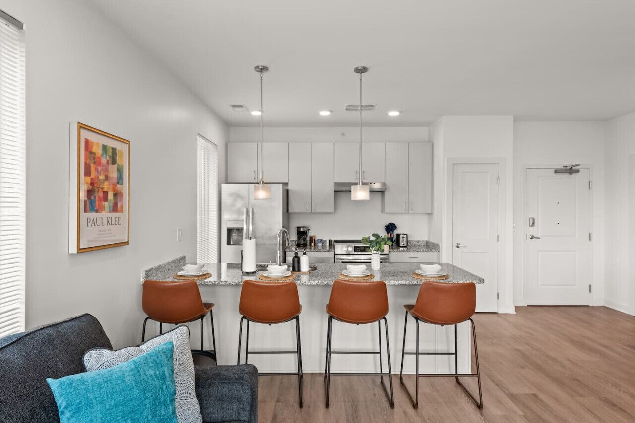 A modern kitchen with white cabinets, a central island with bar stools, pendant lighting, and a framed artwork on the wall.
