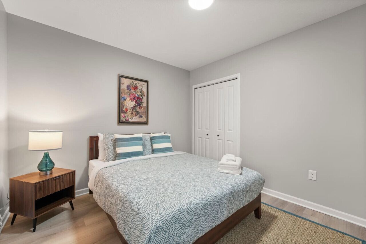 A bedroom with a bed, a nightstand, a lamp, a painting, and a closet.