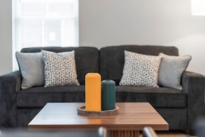 Uma sala de estar moderna com um sofá cinza escuro, almofadas estampadas e uma mesa de centro de madeira com dois objetos cilíndricos em cima.
