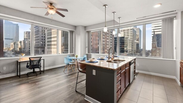 A modern kitchen with a large island, a dining area with blue chairs, and a city view through large windows.