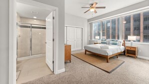 A modern bedroom with a large bed, a walk-in closet, a shower, and a view of the city through large windows.