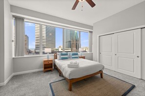 Une chambre avec une grande fenêtre, un lit, une table de chevet, un ventilateur de plafond et une porte.