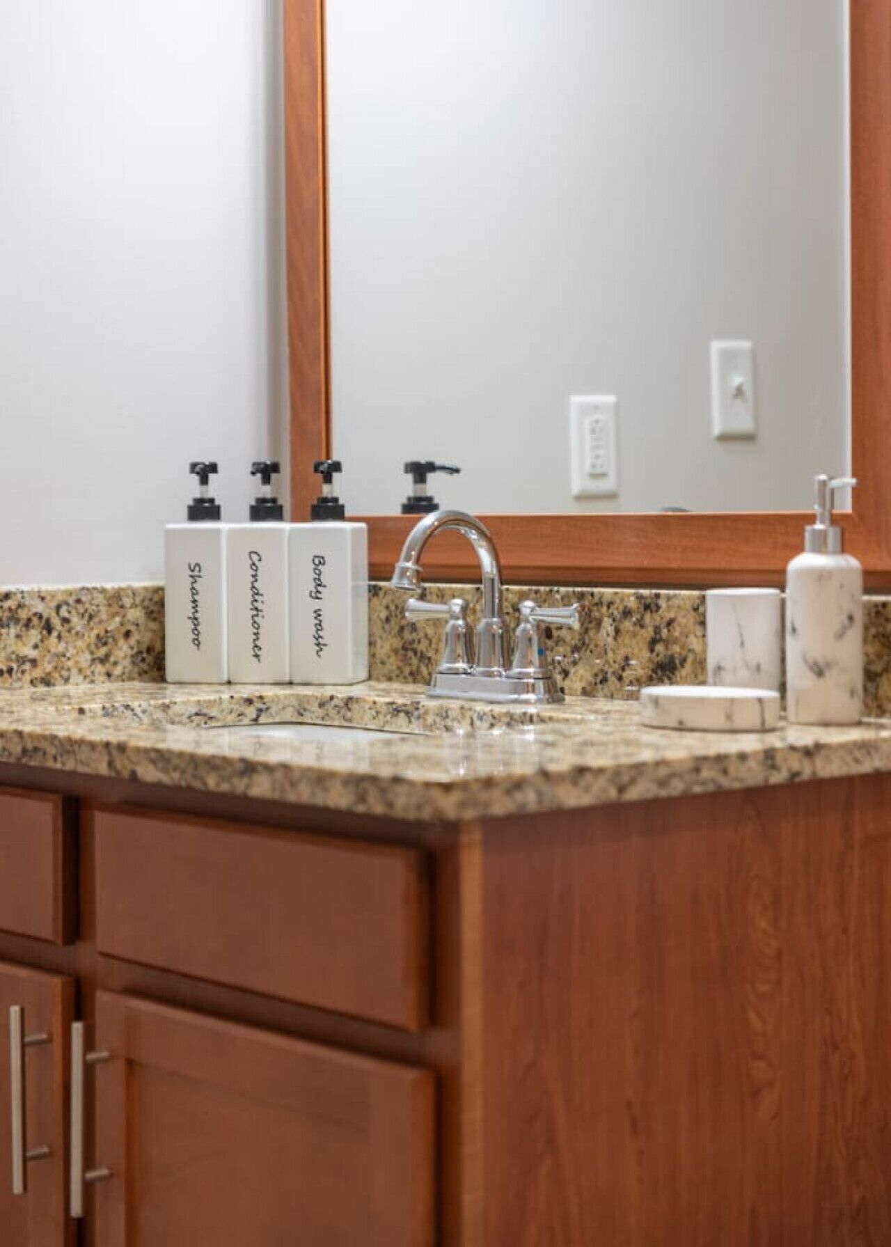 Ein Badezimmer mit einer Granit-Arbeitsfläche, einem Waschbecken mit modernem Wasserhahn und drei weißen Spendern, beschriftet mit 'Shampoo', 'Conditioner' und 'Body Wash'.