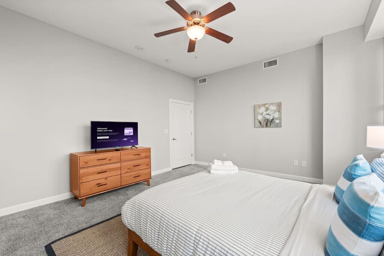 A bedroom with a bed, a dresser, a ceiling fan, a television, and a door.
