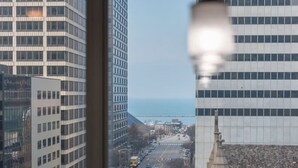 Vista desde una ventana que da a un paisaje urbano con edificios altos, cielo despejado y una masa de agua distante.