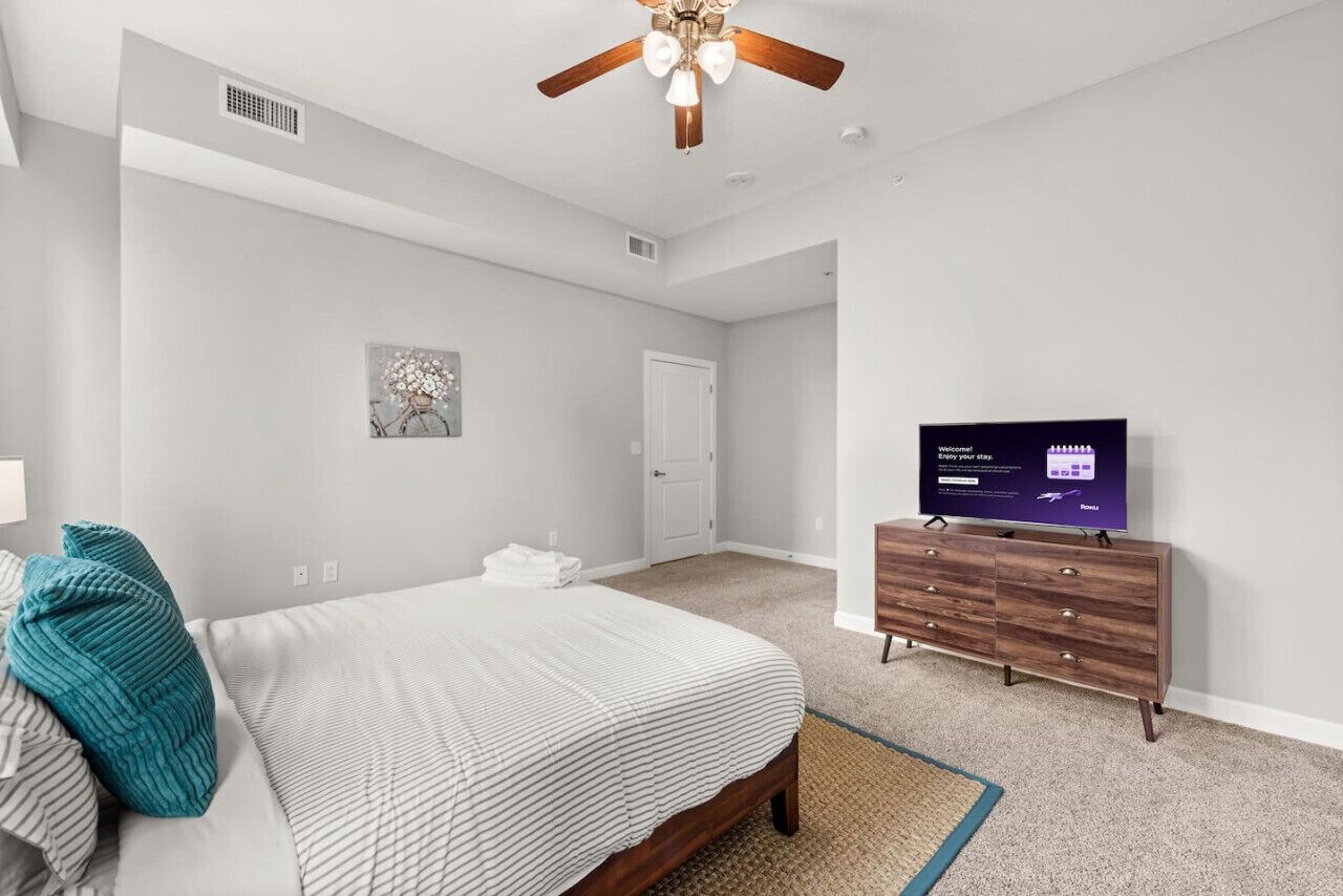 A bedroom with a bed, a dresser, a ceiling fan, a television, and a door.