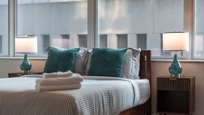 A neatly made bed with pillows, a lamp on each side, and a window with a view of buildings.