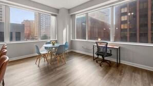 A room with large windows, a dining table with chairs, a desk with a chair, and a lamp.