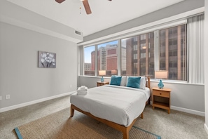 A bedroom with a large window, a bed, a nightstand, and a wall-mounted lamp.