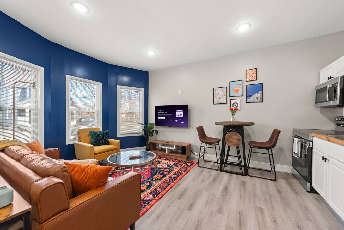 A modern living room with a sofa, coffee table, and a TV. There is a kitchen area with a microwave and cabinets.