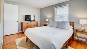 A bedroom with a bed, a dresser, a television, a window with blinds, and a nightstand with a lamp. - Cozy 2BR with Garage | Central North Canton (North Canton)