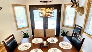 Comedor con una mesa de madera dispuesta para seis comensales, rodeada de sillas tejidas, una araña y una cabeza de animal montada en la pared.