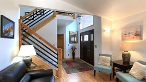 Una sala de estar moderna con una escalera de madera, un sofá de cuero negro, un sillón verde azulado y una mesa de centro de madera.