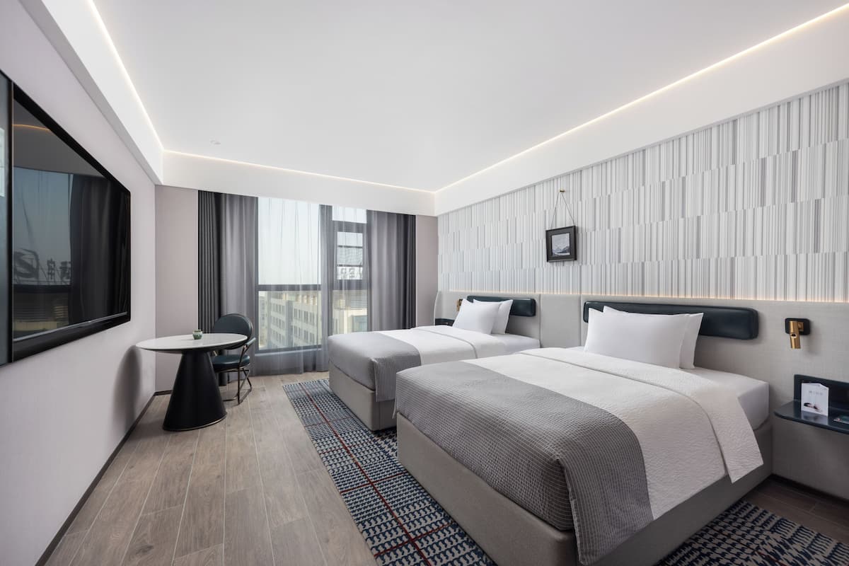 Quarto de hotel moderno com duas camas, uma mesa redonda pequena e vista da cidade através de grandes janelas.