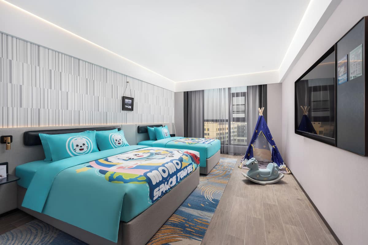 Quarto de hotel moderno com uma cama grande, duas mesas de cabeceira, um espaço de estar e vista para a cidade.