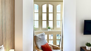 Ein Zimmer mit einer Wand aus Holzlatten, einem Sessel mit Kissen, einem kleinen Tisch mit Pflanze und einem großen Fenster mit vielen Scheiben.
