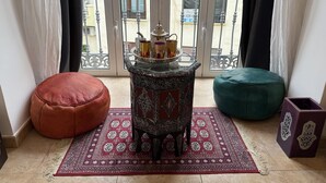 A room with a patterned rug, a small table with a tray of items, a red ottoman, a green pouf, and a decorative box.