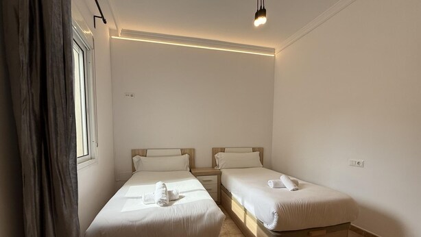 A clean, modern room with two single beds, a wooden nightstand, and a window with curtains.