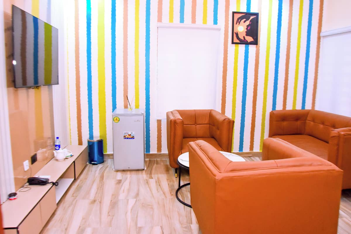 Une pièce avec un papier peint à rayures, deux canapés orange, une petite table, un téléviseur, un réfrigérateur et une poubelle.