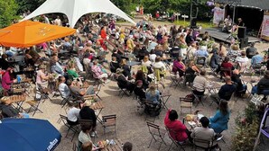 Et udendørs arrangement med folk siddende ved borde under parasoller, en scene og et hvidt telt i baggrunden.