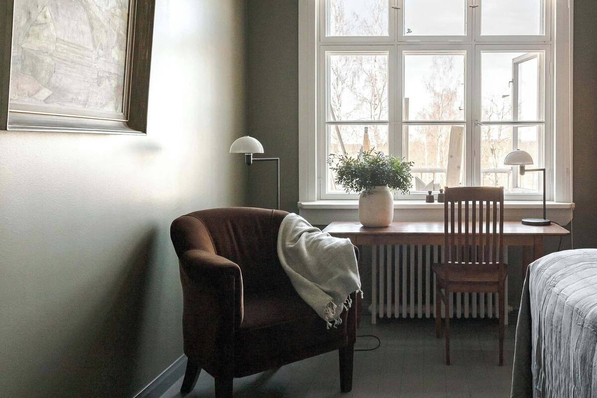 A room with a large window, a radiator, a chair, a table, and a bed.