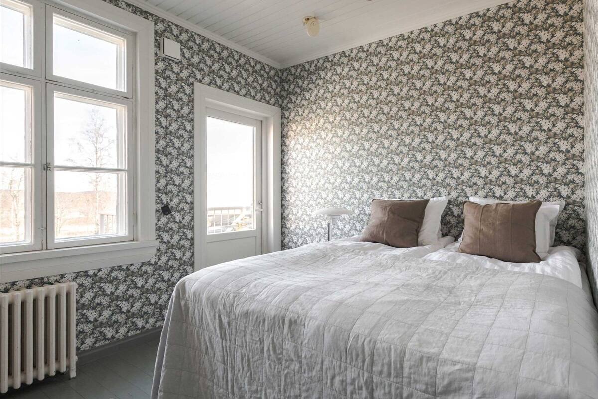 A bedroom with a bed, two windows, a radiator, and floral wallpaper.