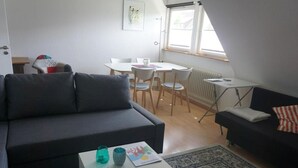 A modern living room with a dark grey sofa, a white coffee table, and a black sofa with a striped pillow. There is a dining area with a white table and chairs, and a window with blinds. - Apartment 'Kirsch-kern' with Balcony and Wi-Fi (Mössingen)