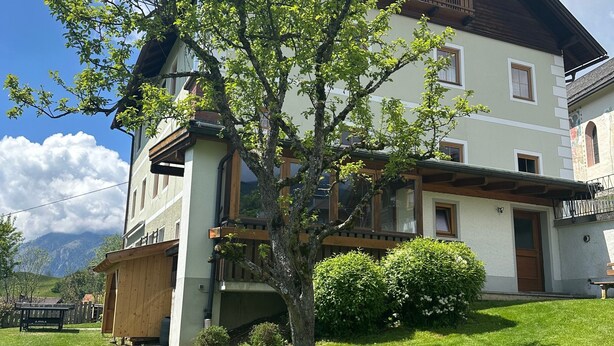 Un edificio de dos pisos con techo inclinado, una terraza de madera y un pequeño árbol en el jardín delantero.