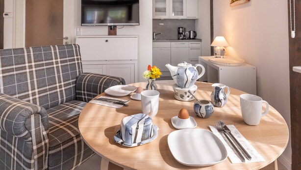 Una sala de estar moderna con un sofá a cuadros, una mesa de comedor dispuesta con platos, tazas y cubiertos, y una cocina al fondo.