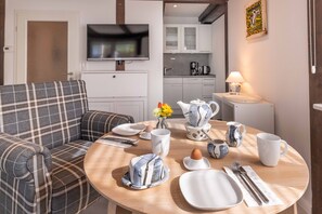Una sala de estar moderna con un sofá a cuadros, una mesa de comedor dispuesta con platos, tazas y cubiertos, y una cocina al fondo.